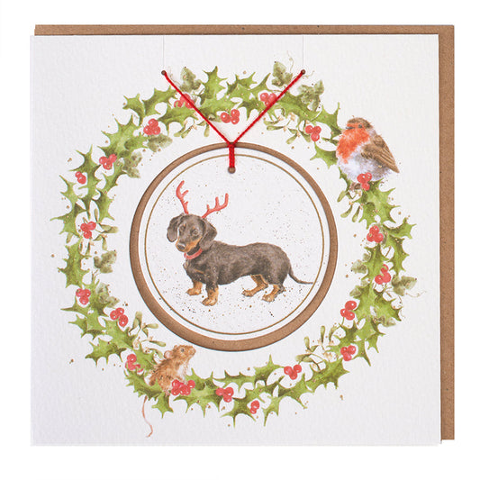 Wrendale Designs Christmas card with foiled, detachable decoration featuring Hannah Dale's artwork of a Dachshund reluctantly wearing Reindeer antlers and titled <i>Dachshund through the Snow</i>